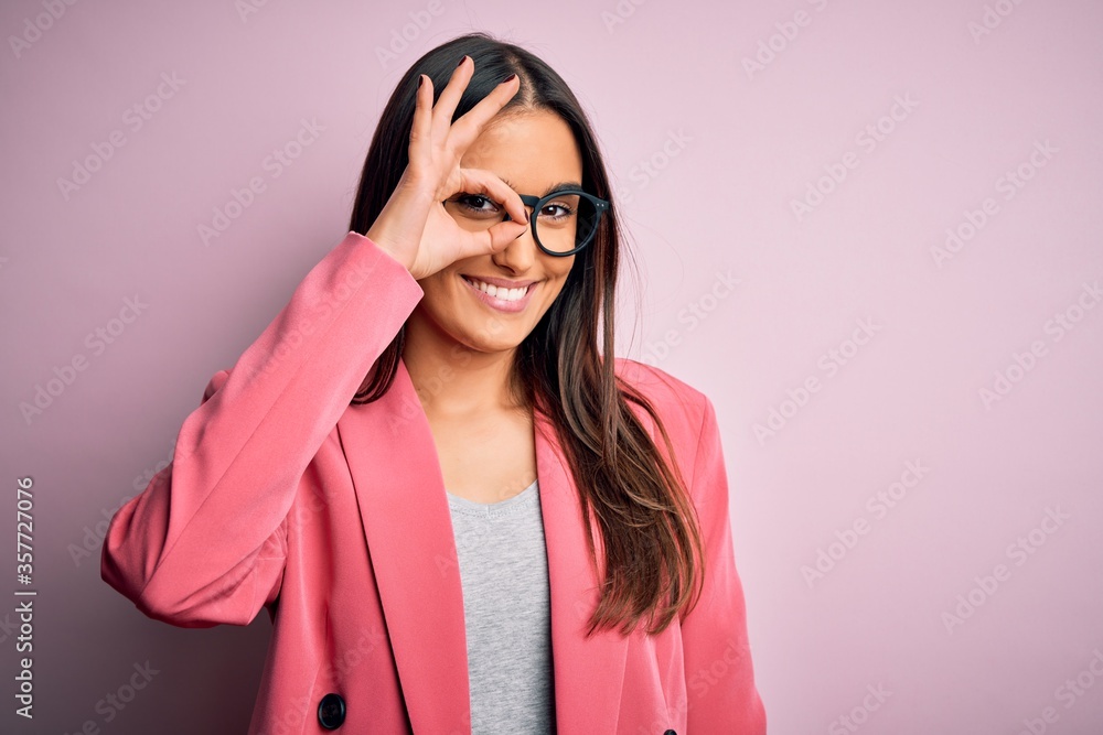 Young beautiful brunette businesswoman wearing jacket and glasses over pink background doing ok gesture with hand smiling, eye looking through fingers with happy face.