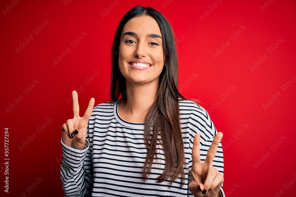 Young beautiful brunette woman wearing casual striped t-shirt over red background smiling looking to the camera showing fingers doing victory sign. Number two. Young beautiful brunette woman wearing casual striped t-shirt over red background smiling looking to the camera showing fingers doing victory sign. Number two.