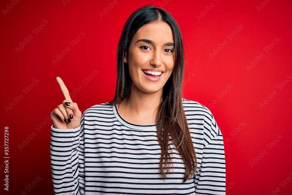 Young beautiful brunette woman wearing casual striped t-shirt over red background showing and pointing up with finger number one while smiling confident and happy. Young beautiful brunette woman wearing casual striped t-shirt over red background showing and pointing up with finger number one while smiling confident and happy.