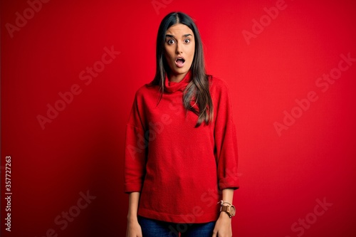 Young beautiful brunette woman wearing casual sweater over isolated red background In shock face, looking skeptical and sarcastic, surprised with open mouth