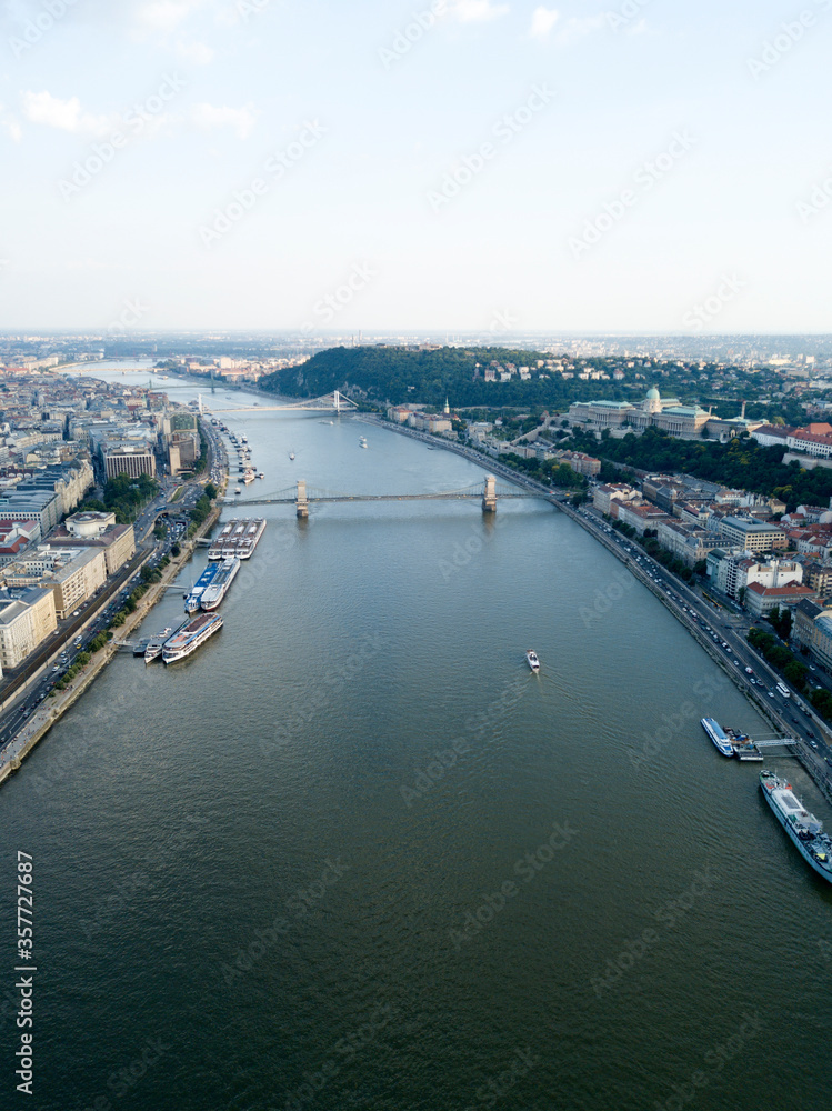 Aerial view of Széchenyi Chain Bridge over Danube river near Parliament palace in Budapest city, Hungary. Cars drive on bridge, road, boats floating on river, ships moored near riverside. Historic cen