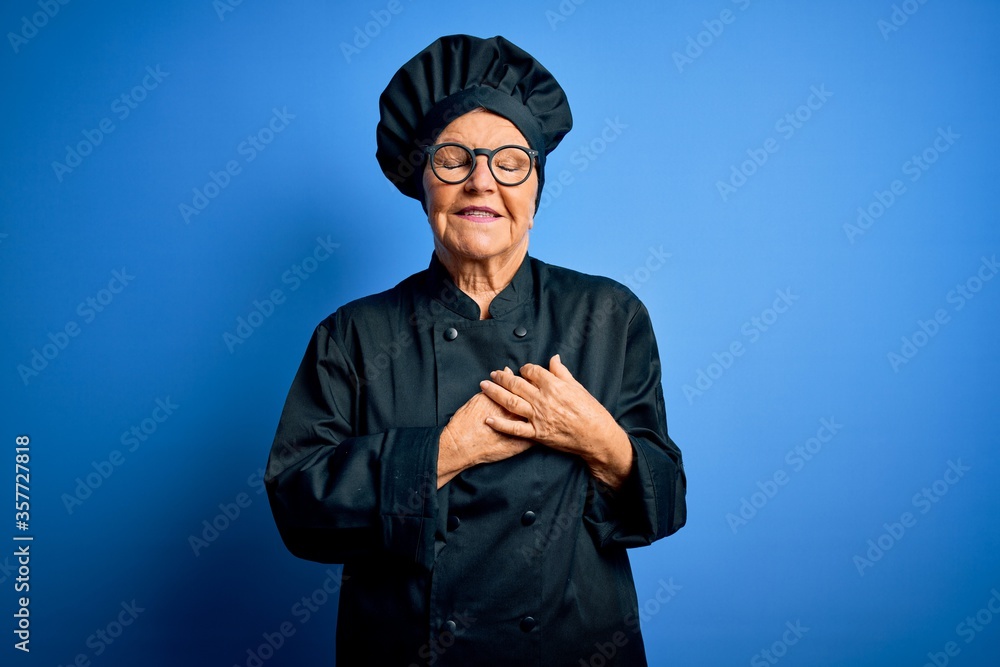 Senior beautiful grey-haired chef woman wearing cooker uniform and hat over blue background smiling with hands on chest with closed eyes and grateful gesture on face. Health concept. Senior beautiful grey-haired chef woman wearing cooker uniform and hat over blue background smiling with hands on chest with closed eyes and grateful gesture on face. Health concept.