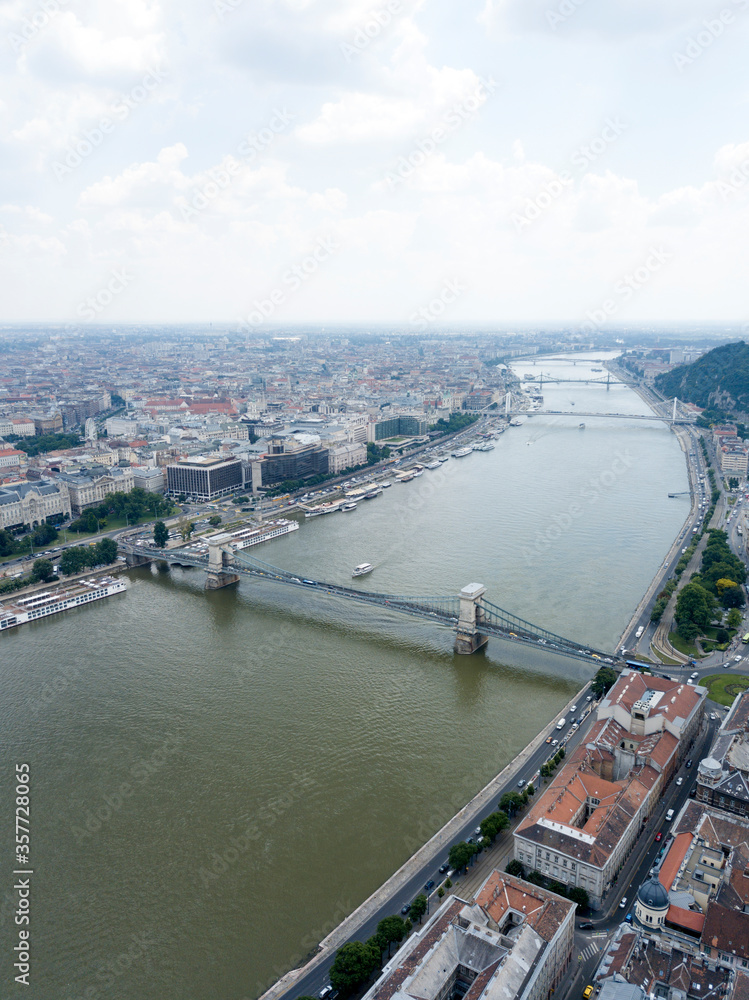 Aerial view of Széchenyi Chain Bridge over Danube river near Parliament palace in Budapest city, Hungary. Cars drive on bridge, road, boats floating on river, ships moored near riverside. Historic cen