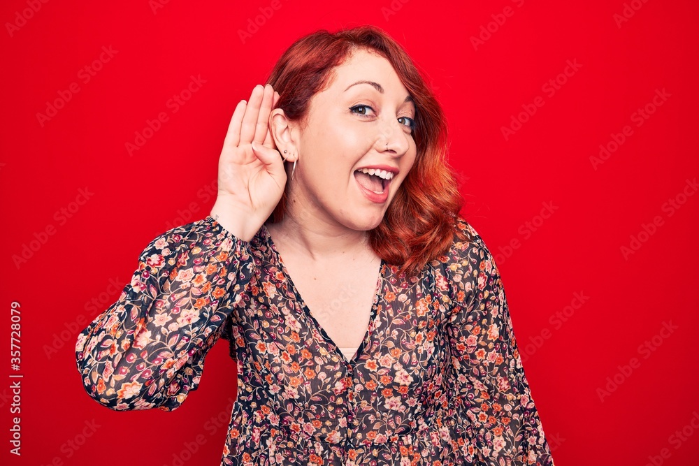 Young beautiful redhead woman wearing floral casual dress standing over red background smiling with hand over ear listening and hearing to rumor or gossip. Deafness concept. Young beautiful redhead woman wearing floral casual dress standing over red background smiling with hand over ear listening and hearing to rumor or gossip. Deafness concept.