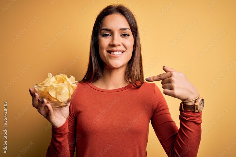 Young beautiful brunette woman holding bowl with chips potatoes over yellow background with surprise face pointing finger to himself