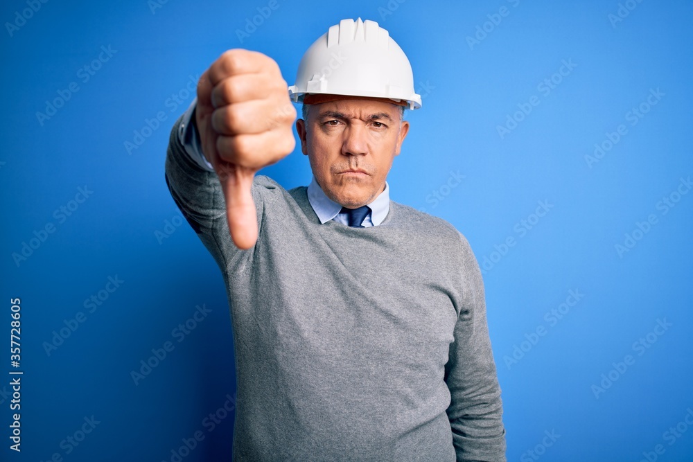Middle age handsome grey-haired engineer man wearing safety helmet over blue background looking unhappy and angry showing rejection and negative with thumbs down gesture. Bad expression. Middle age handsome grey-haired engineer man wearing safety helmet over blue background looking unhappy and angry showing rejection and negative with thumbs down gesture. Bad expression.
