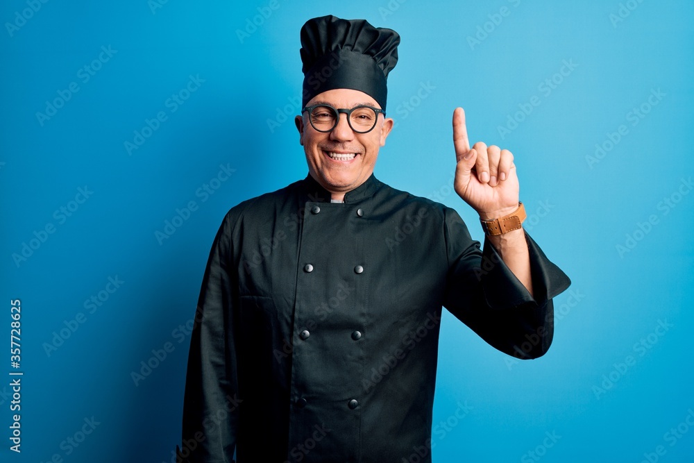 Middle age handsome grey-haired chef man wearing cooker uniform and hat showing and pointing up with finger number one while smiling confident and happy. Middle age handsome grey-haired chef man wearing cooker uniform and hat showing and pointing up with finger number one while smiling confident and happy.