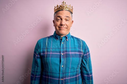 Middle age handsome grey-haired man wearing golden crown of king over pink background with a happy and cool smile on face. Lucky person.