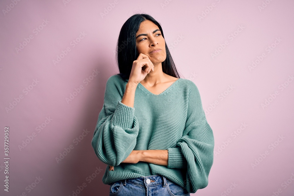 Young beautiful hispanic fashion woman wearing green sweater over pink background with hand on chin thinking about question, pensive expression. Smiling with thoughtful face. Doubt concept. Young beautiful hispanic fashion woman wearing green sweater over pink background with hand on chin thinking about question, pensive expression. Smiling with thoughtful face. Doubt concept.