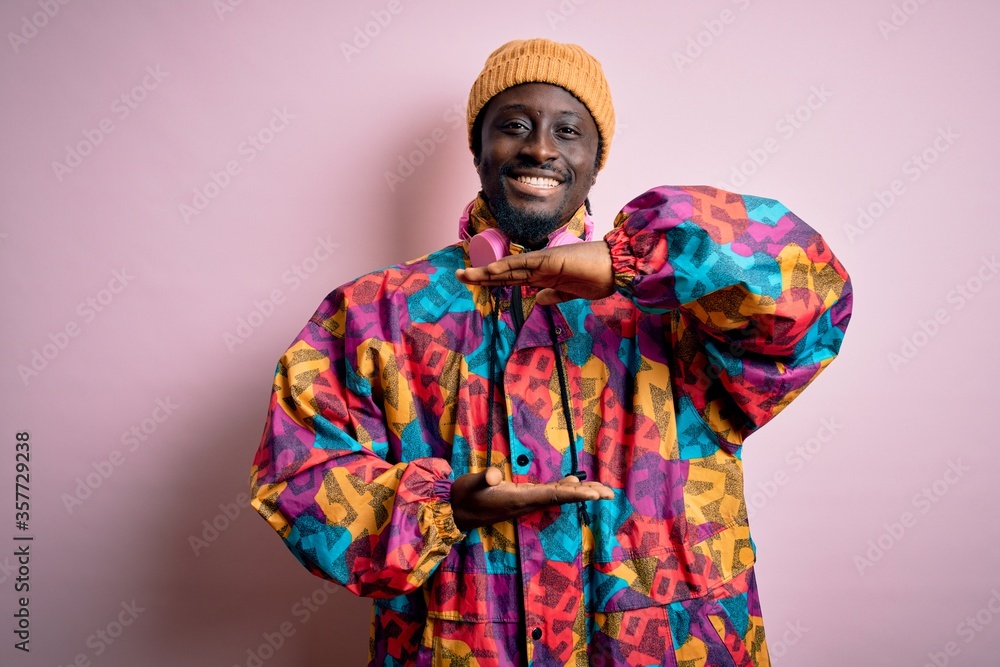 Obraz premium Young handsome african american man wearing colorful coat and cap over pink background gesturing with hands showing big and large size sign, measure symbol. Smiling looking at the camera. Measuring