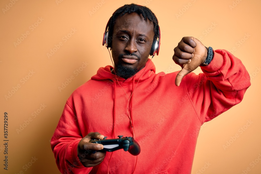 Young african american gamer man playing video game using joystick and headphones with angry face, negative sign showing dislike with thumbs down, rejection concept Young african american gamer man playing video game using joystick and headphones with angry face, negative sign showing dislike with thumbs down, rejection concept