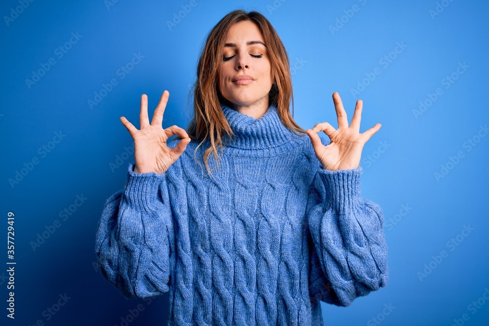 Young beautiful brunette woman wearing casual turtleneck sweater over blue background relax and smiling with eyes closed doing meditation gesture with fingers. Yoga concept. Young beautiful brunette woman wearing casual turtleneck sweater over blue background relax and smiling with eyes closed doing meditation gesture with fingers. Yoga concept.
