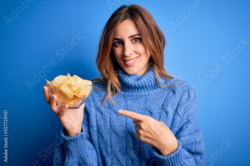 Young beautiful brunette woman holding bowl with potato chips over blue background very happy pointing with hand and finger