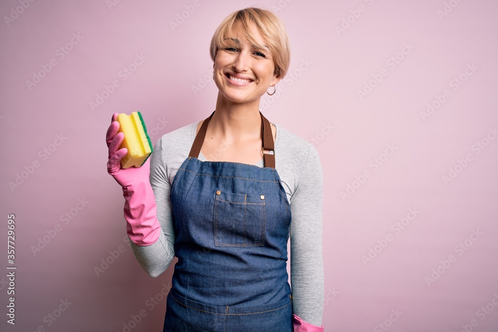 Young blonde cleaner woman with short hair wearing apron and gloves holding scrub scourer with a happy face standing and smiling with a confident smile showing teeth Young blonde cleaner woman with short hair wearing apron and gloves holding scrub scourer with a happy face standing and smiling with a confident smile showing teeth