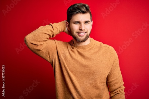 Young blond man with beard and blue eyes wearing casual sweater over red background confuse and wonder about question. Uncertain with doubt, thinking with hand on head. Pensive concept. © Krakenimages.com