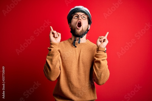 Young blond cyclist man with beard and blue eyes wearing bike helmet and glasses amazed and surprised looking up and pointing with fingers and raised arms.