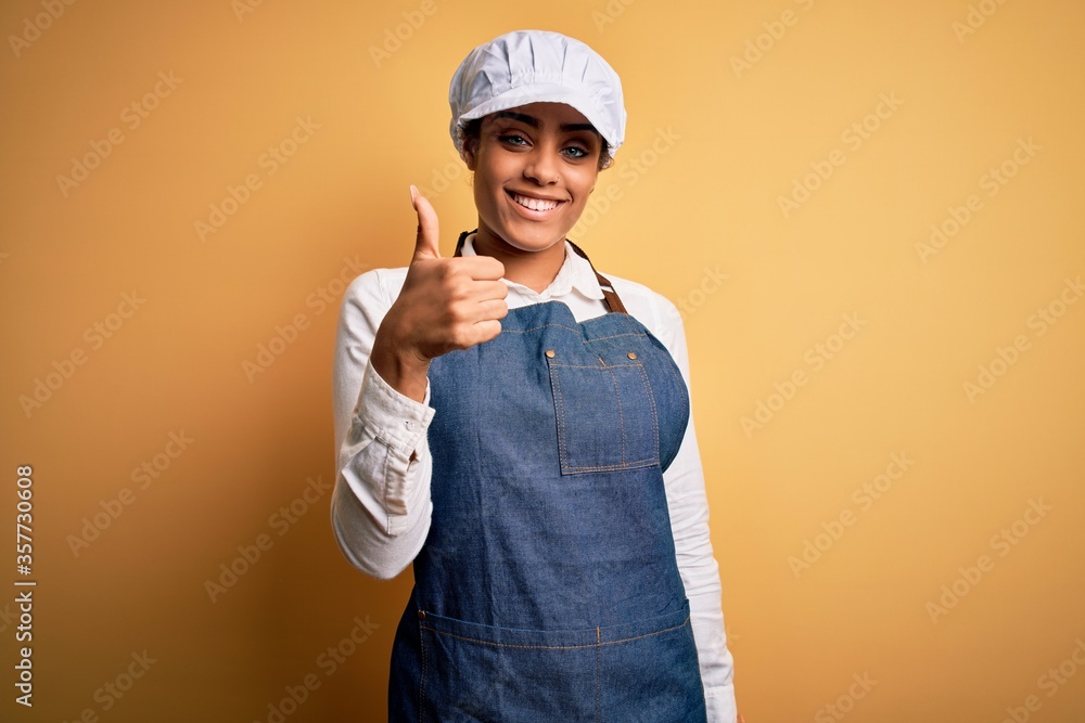 Young african american afro baker woman wearing apron and cap over yellow background doing happy thumbs up gesture with hand. Approving expression looking at the camera showing success.