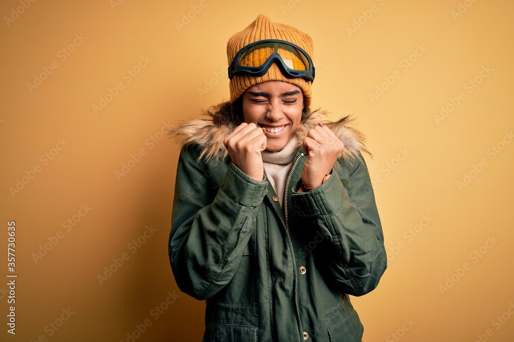 Young african american afro skier girl wearing snow sportswear and ski goggles excited for success with arms raised and eyes closed celebrating victory smiling. Winner concept. Young african american afro skier girl wearing snow sportswear and ski goggles excited for success with arms raised and eyes closed celebrating victory smiling. Winner concept.