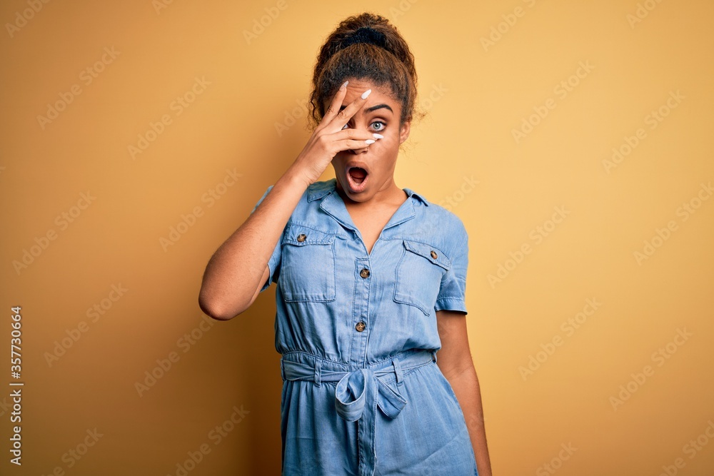 Young beautiful african american girl wearing denim dress standing over yellow background peeking in shock covering face and eyes with hand, looking through fingers with embarrassed expression. Young beautiful african american girl wearing denim dress standing over yellow background peeking in shock covering face and eyes with hand, looking through fingers with embarrassed expression.