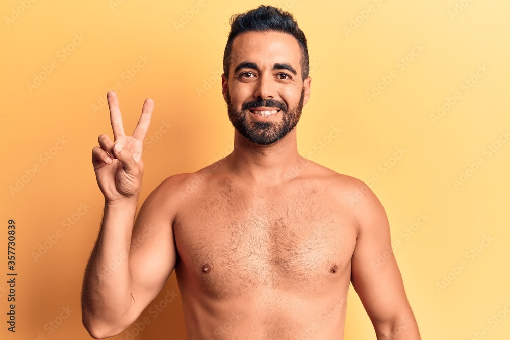 Young hispanic man standing shirtless smiling with happy face winking at the camera doing victory sign. number two.