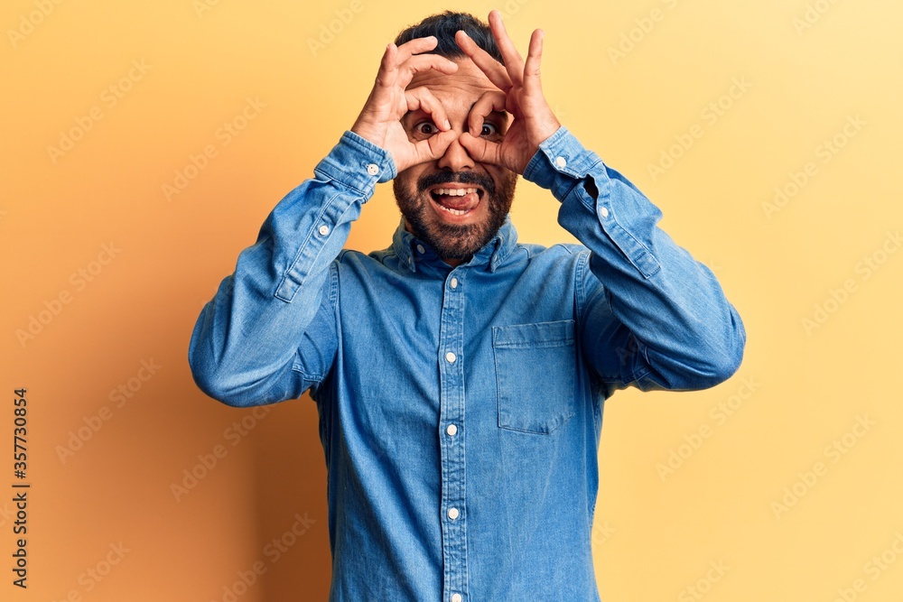 Young hispanic man wearing casual clothes doing ok gesture like binoculars sticking tongue out, eyes looking through fingers. crazy expression. Young hispanic man wearing casual clothes doing ok gesture like binoculars sticking tongue out, eyes looking through fingers. crazy expression.