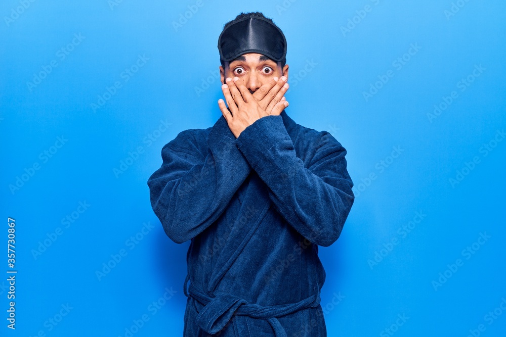 Young hispanic man wearing sleep mask and robe shocked covering mouth with hands for mistake. secret concept. Young hispanic man wearing sleep mask and robe shocked covering mouth with hands for mistake. secret concept.