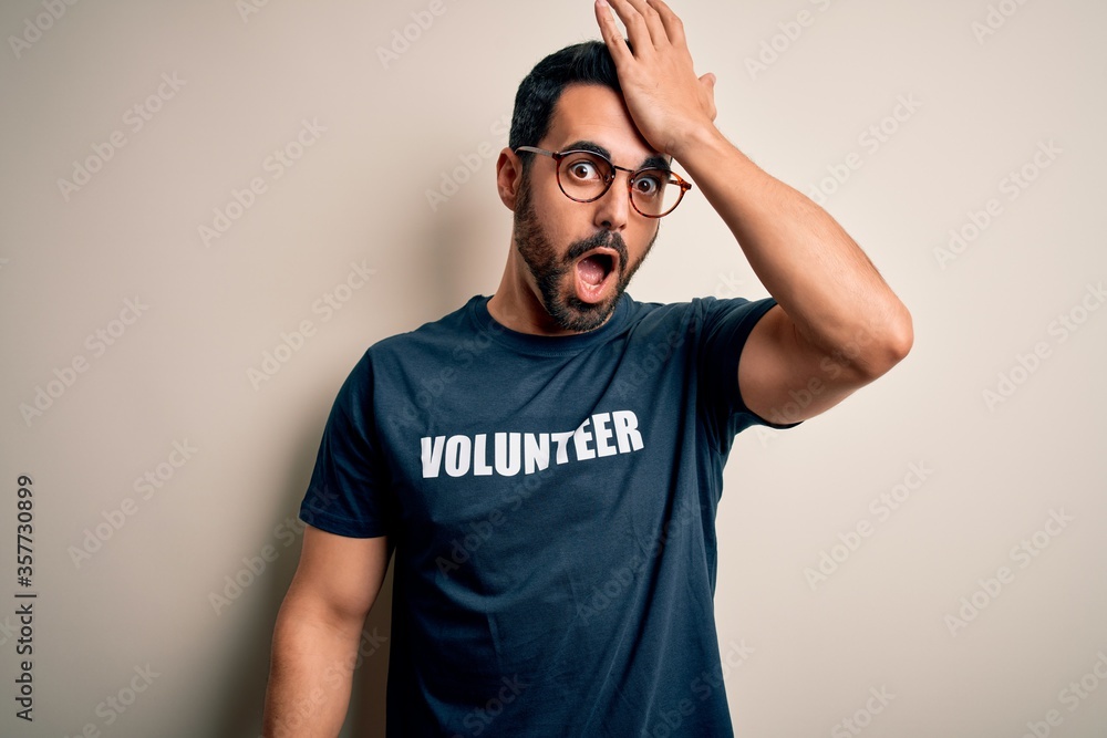 Handsome man with beard wearing t-shirt with volunteer message over white background surprised with hand on head for mistake, remember error. Forgot, bad memory concept. Handsome man with beard wearing t-shirt with volunteer message over white background surprised with hand on head for mistake, remember error. Forgot, bad memory concept.