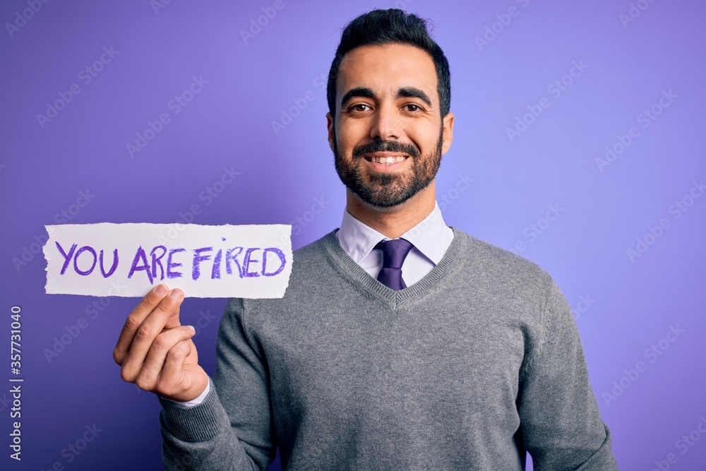 Young handsome businessman with beard holding paper with you are fired message with a happy face standing and smiling with a confident smile showing teeth Young handsome businessman with beard holding paper with you are fired message with a happy face standing and smiling with a confident smile showing teeth