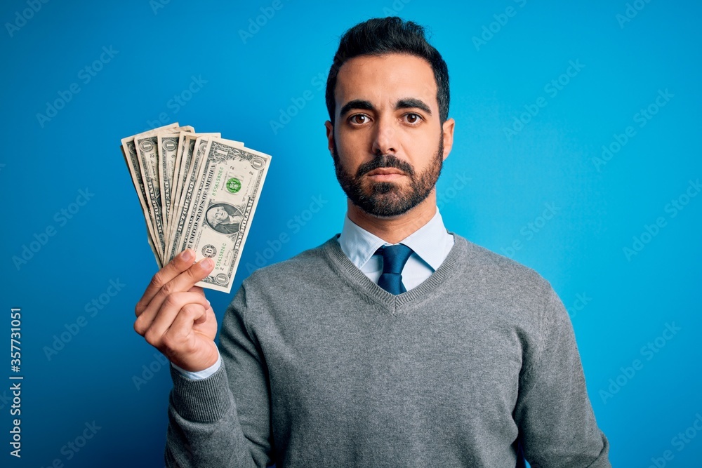 Young handsome man with beard holding bunch of dollars banknotes over blue background with a confident expression on smart face thinking serious Young handsome man with beard holding bunch of dollars banknotes over blue background with a confident expression on smart face thinking serious