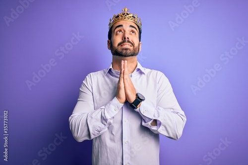 Young handsome man with beard wearing golden crown of king over purple background begging and praying with hands together with hope expression on face very emotional and worried. Begging. © Krakenimages.com