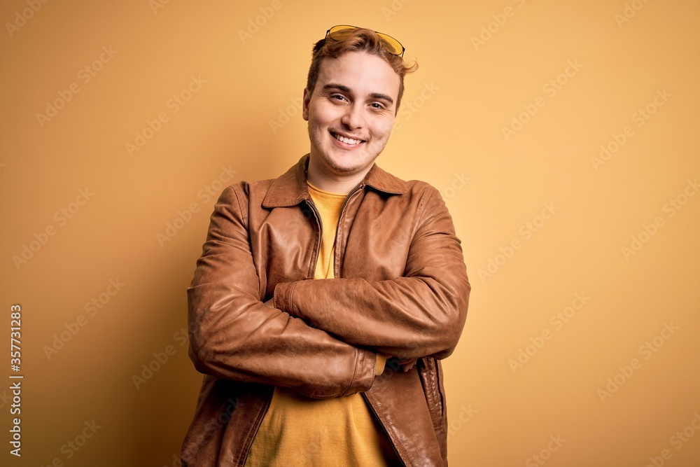 Young handsome redhead man wearing casual leather jacket over isolated yellow background happy face smiling with crossed arms looking at the camera. Positive person.