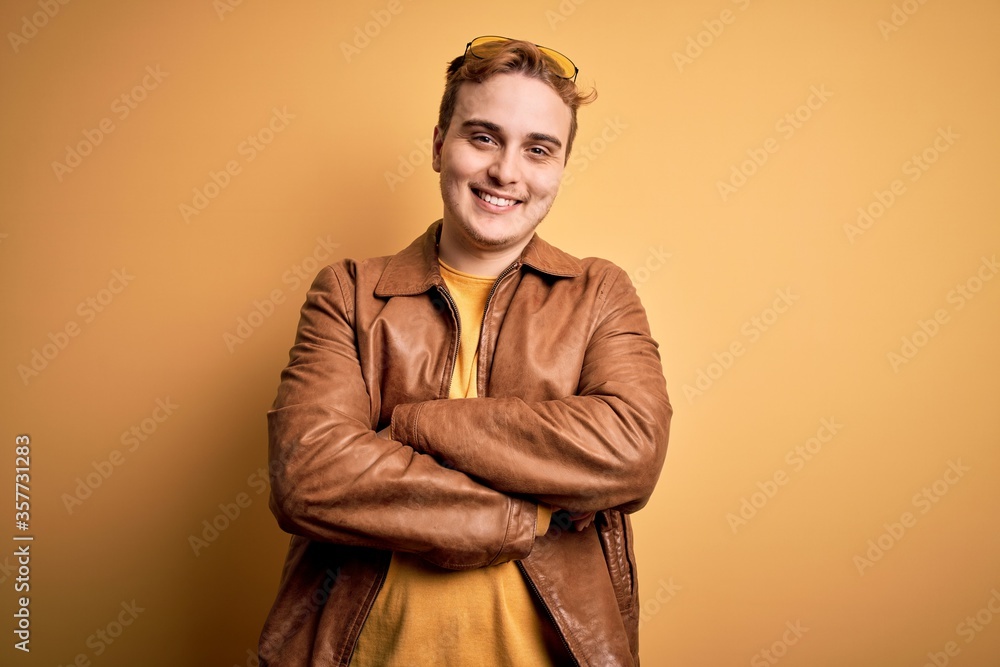 Young handsome redhead man wearing casual leather jacket over isolated yellow background happy face smiling with crossed arms looking at the camera. Positive person. Young handsome redhead man wearing casual leather jacket over isolated yellow background happy face smiling with crossed arms looking at the camera. Positive person.