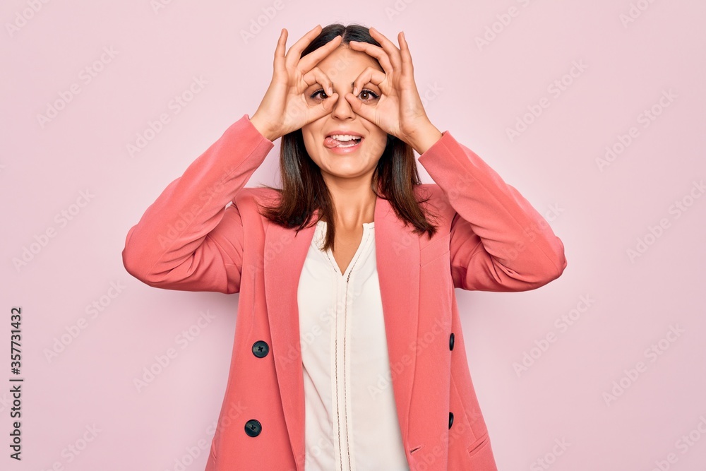 Young beautiful brunette businesswoman wearing elegant jacket over isolated pink background doing ok gesture like binoculars sticking tongue out, eyes looking through fingers. Crazy expression. Young beautiful brunette businesswoman wearing elegant jacket over isolated pink background doing ok gesture like binoculars sticking tongue out, eyes looking through fingers. Crazy expression.