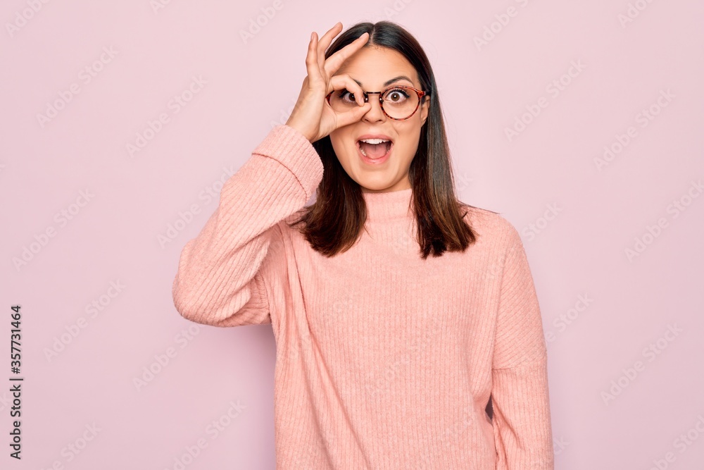 Young beautiful brunette woman wearing casual sweater and glasses over pink background doing ok gesture with hand smiling, eye looking through fingers with happy face.