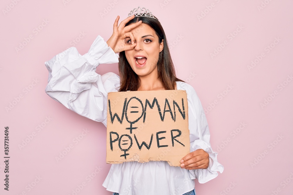 Girl wearing princess tiara asking for women rights holding banner with woman power message smiling happy doing ok sign with hand on eye looking through fingers Girl wearing princess tiara asking for women rights holding banner with woman power message smiling happy doing ok sign with hand on eye looking through fingers