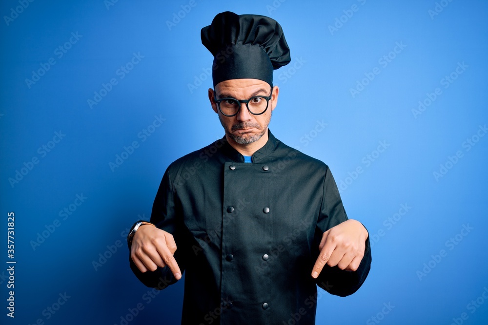 Young handsome chef man wearing cooker uniform and hat over isolated blue background Pointing down looking sad and upset, indicating direction with fingers, unhappy and depressed. Young handsome chef man wearing cooker uniform and hat over isolated blue background Pointing down looking sad and upset, indicating direction with fingers, unhappy and depressed.
