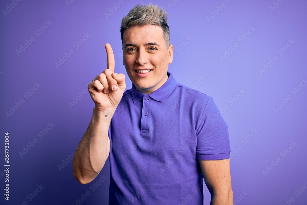 Young handsome modern man wearing casual purple t-shirt over isolated background showing and pointing up with finger number one while smiling confident and happy.