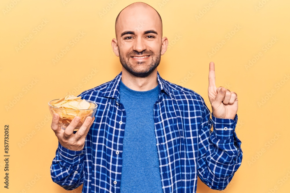 Young handsome bald man holding bowl with potato chips smiling with an idea or question pointing finger with happy face, number one Young handsome bald man holding bowl with potato chips smiling with an idea or question pointing finger with happy face, number one