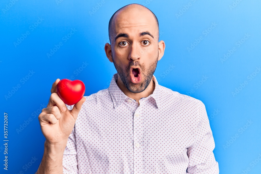Young handsome bald man holding heart scared and amazed with open mouth for surprise, disbelief face Young handsome bald man holding heart scared and amazed with open mouth for surprise, disbelief face