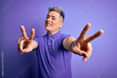 Young handsome modern man wearing casual purple t-shirt over isolated background smiling looking to the camera showing fingers doing victory sign. Number two.