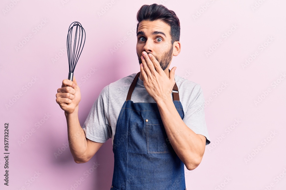 Young handsome man with beard wearing apron holding whisk covering mouth with hand, shocked and afraid for mistake. surprised expression Young handsome man with beard wearing apron holding whisk covering mouth with hand, shocked and afraid for mistake. surprised expression