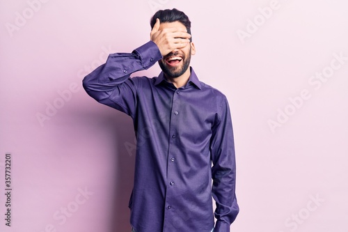Young handsome man with beard wearing casual shirt smiling and laughing with hand on face covering eyes for surprise. blind concept.