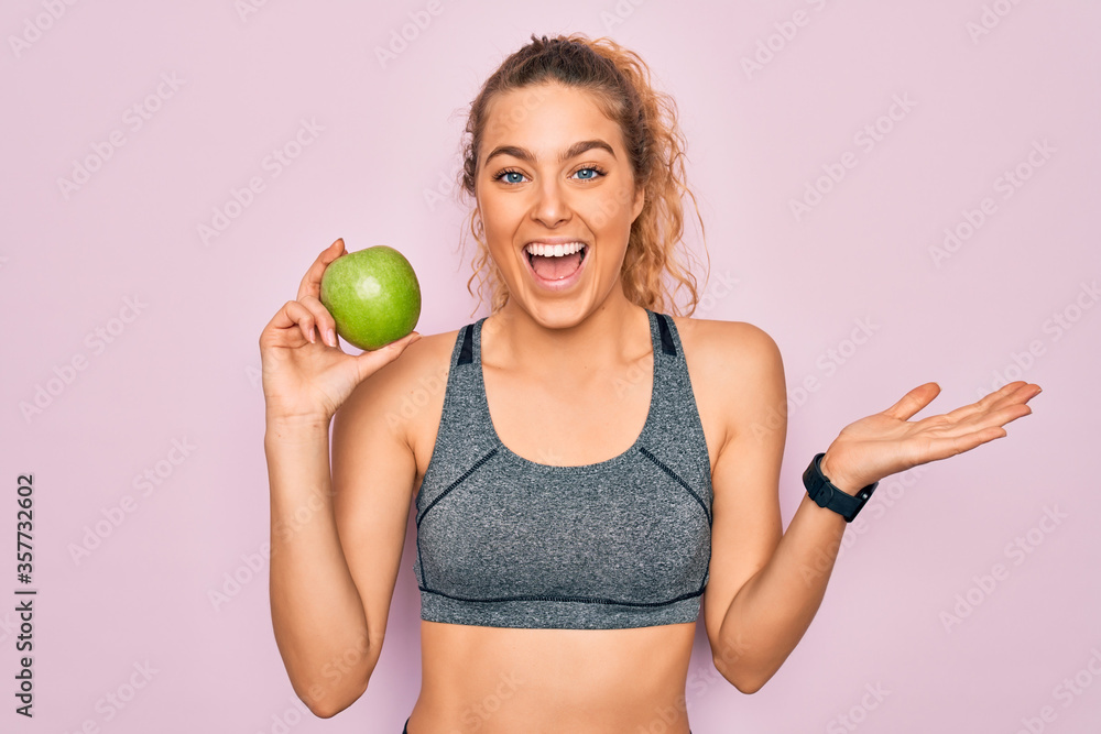 Young beautiful blonde sporty woman with blue eyes holding healthy green apple fruit very happy and excited, winner expression celebrating victory screaming with big smile and raised hands Young beautiful blonde sporty woman with blue eyes holding healthy green apple fruit very happy and excited, winner expression celebrating victory screaming with big smile and raised hands