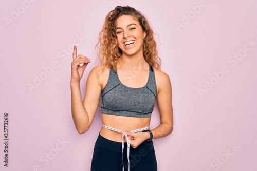 Young blonde sporty woman with blue eyes controlling weight using tape measure on waist surprised with an idea or question pointing finger with happy face, number one photo