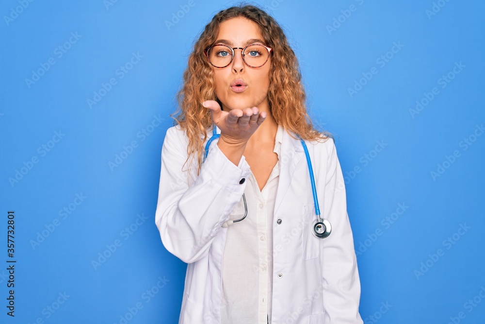 Young beautiful blonde doctor woman with blue eyes wearing coat and stethoscope looking at the camera blowing a kiss with hand on air being lovely and sexy. Love expression. Young beautiful blonde doctor woman with blue eyes wearing coat and stethoscope looking at the camera blowing a kiss with hand on air being lovely and sexy. Love expression.