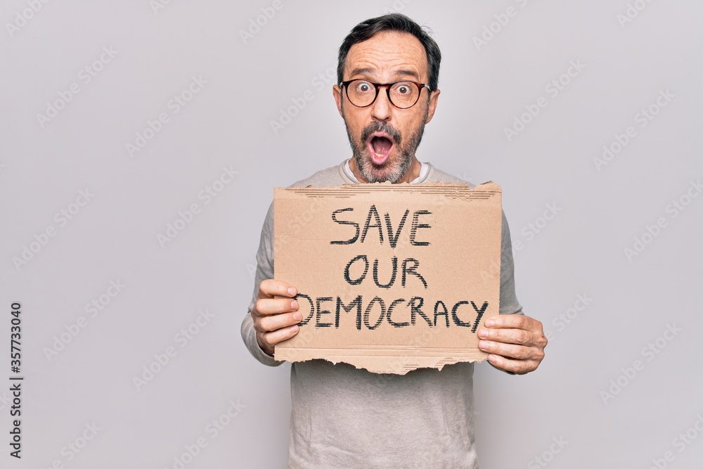 Middle age man holding banner with saver our democracy message over white background scared and amazed with open mouth for surprise, disbelief face Middle age man holding banner with saver our democracy message over white background scared and amazed with open mouth for surprise, disbelief face