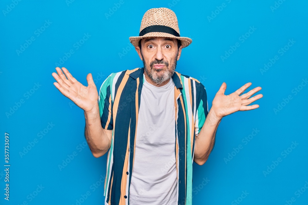 Middle age handsome tourist man on vacation wearing shirt and hat over blue background clueless and confused with open arms, no idea and doubtful face. Middle age handsome tourist man on vacation wearing shirt and hat over blue background clueless and confused with open arms, no idea and doubtful face.