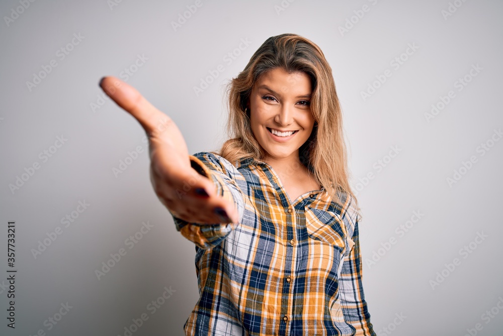 Young beautiful blonde woman wearing casual shirt standing over isolated white background smiling friendly offering handshake as greeting and welcoming. Successful business. Young beautiful blonde woman wearing casual shirt standing over isolated white background smiling friendly offering handshake as greeting and welcoming. Successful business.