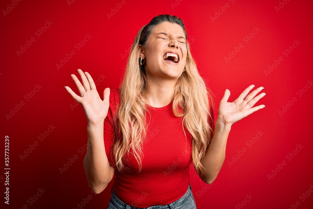 Young beautiful blonde woman wearing casual t-shirt standing over isolated red background celebrating mad and crazy for success with arms raised and closed eyes screaming excited. Winner concept Young beautiful blonde woman wearing casual t-shirt standing over isolated red background celebrating mad and crazy for success with arms raised and closed eyes screaming excited. Winner concept