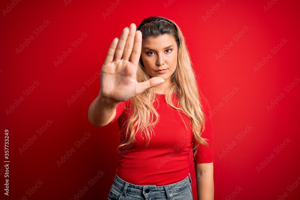 Young beautiful blonde woman wearing casual t-shirt standing over isolated red background doing stop sing with palm of the hand. Warning expression with negative and serious gesture on the face. Young beautiful blonde woman wearing casual t-shirt standing over isolated red background doing stop sing with palm of the hand. Warning expression with negative and serious gesture on the face.